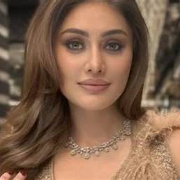 Portrait of a young Indian woman with wavy brown hair, soft glam makeup, beige feather-textured dress and diamond necklace indoors.